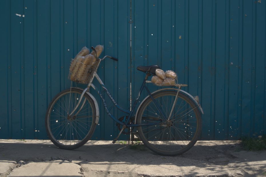 Bicicletta con pane nel portapacchi, poggiata su un cancello crivellato di colpi da arma da fuoco.